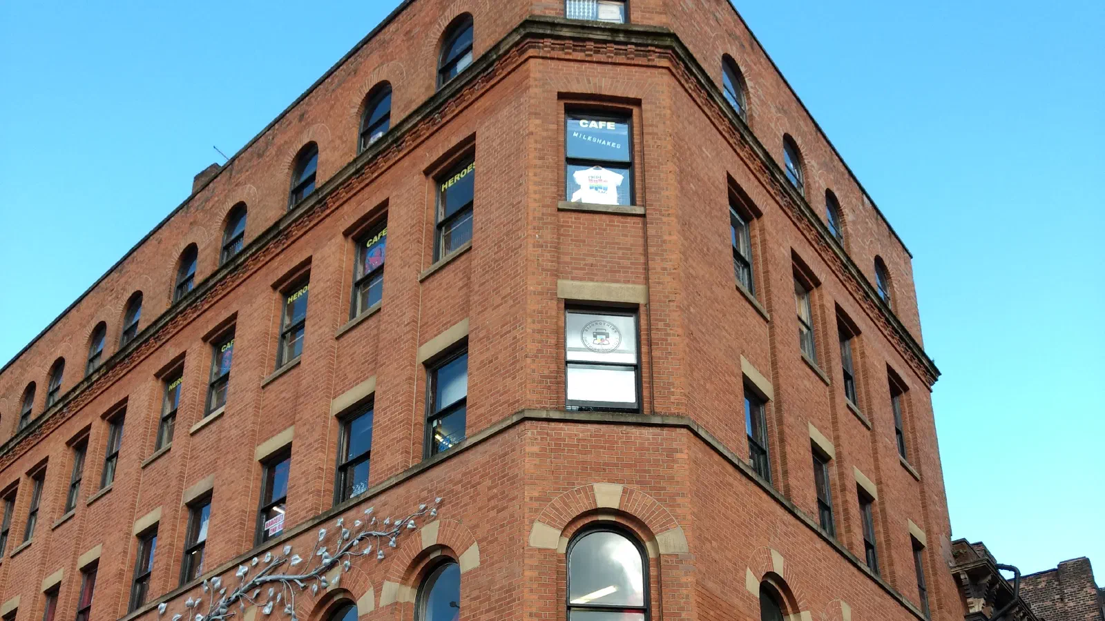 Red brick Victorian corner building with rows of arched windows, poster-covered ground-floor shopfronts and an arched doorway, under a clear blue sky. — Afflecks, Manchester