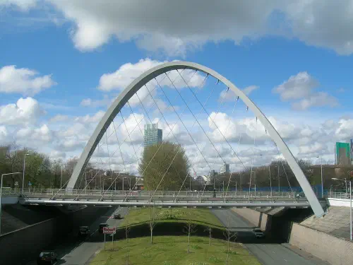 A cable-stayed bridge spans a grassy park under a blue sky with white clouds and distant buildings visible through its arch.