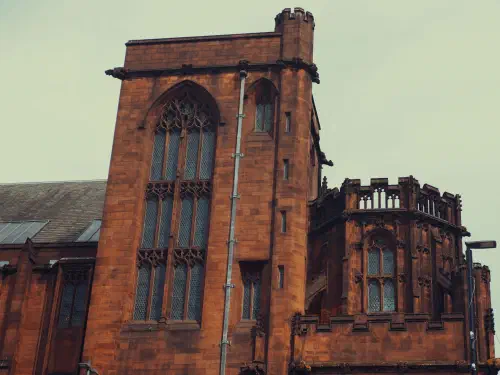 A red sandstone Gothic building with tall arched windows, a metal drainpipe, and an overcast sky.