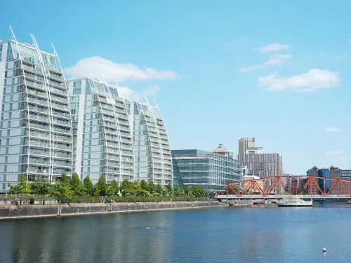 Modern glass skyscrapers line a riverfront park under blue sky and white clouds on sunny day.